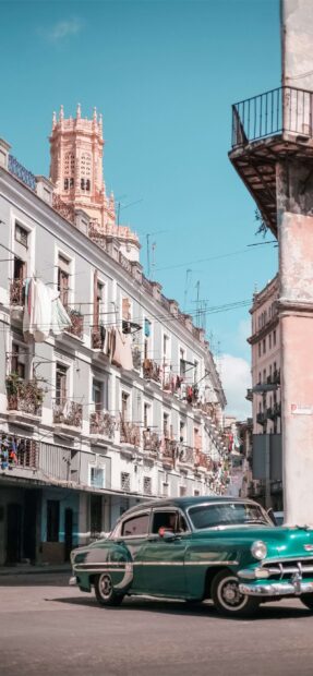 A classic vintage car drives down a bright street in Havana Cuba under a clear blue sky, 2K Mobile Wallpaper