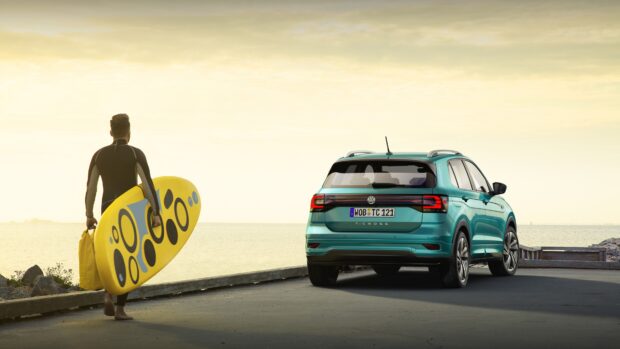 A man carrying a surfboard walks toward a Volkswagen T Cross near the ocean, 4K Desktop Wallpaper