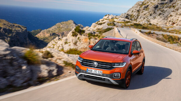 A bright orange Volkswagen T Cross is driving on a winding road near rocky cliffs and the sea, 4K Desktop Wallpaper