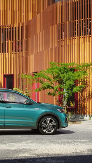 A teal Volkswagen T Cross parked beside a modern building with orange vertical panels and greenery, 2K Mobile Wallpaper