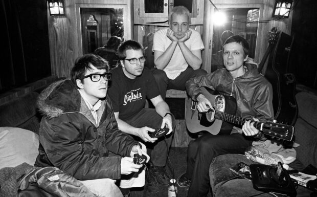 The Weezer band members are sitting together playing video games and guitar inside a room, 2K Desktop Wallpaper