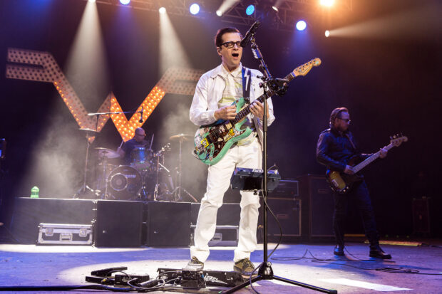 The band Weezer performs on stage with instruments during a concert in front of a large lit logo, HD Desktop Wallpaper