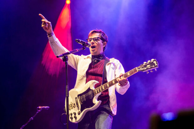 A guitarist from Weezer performing live with a white electric guitar and vibrant stage lights, 2K Desktop Wallpaper