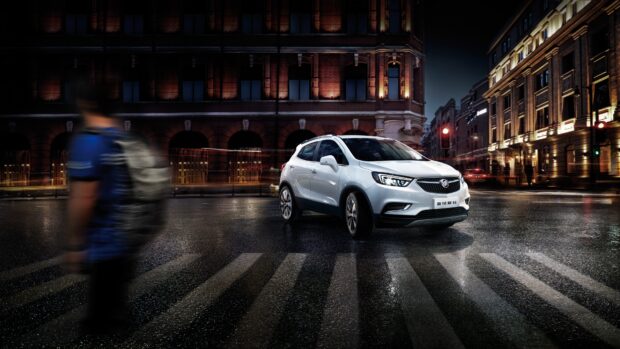 A white Buick Encore drives through a wet city crosswalk with blurred pedestrians nearby, 4K Desktop Wallpaper