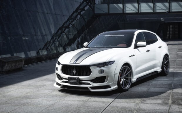 A white Maserati Levante with black racing stripes is parked in front of a modern glass building, 2K Desktop Wallpaper