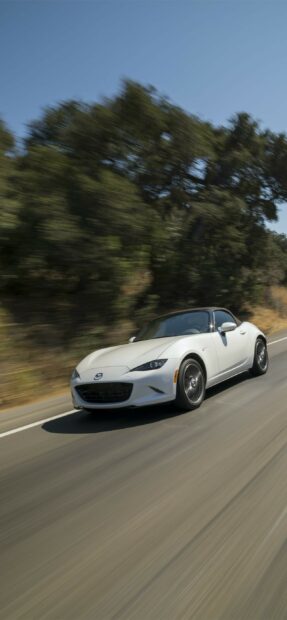 A white Mazda MX 5 Miata is driving fast on a road surrounded by trees, 2K Phone Wallpaper