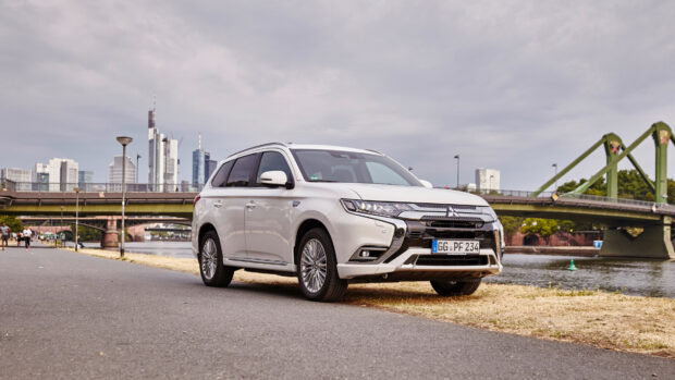 A white Mitsubishi Outlander is parked on a riverside path with city buildings and a green bridge in the background, 4K Desktop Wallpaper
