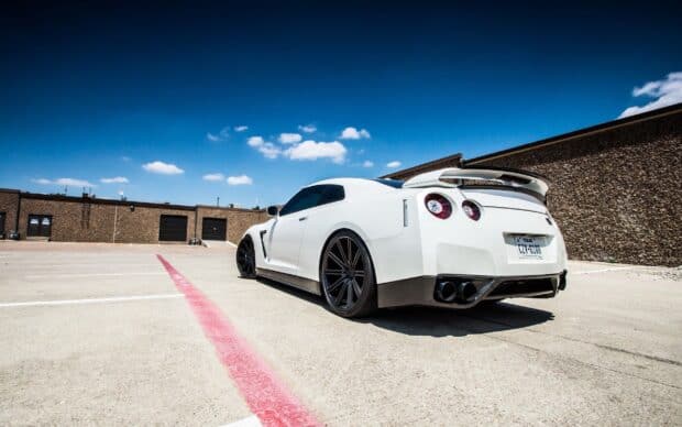 A white Nissan GT R with black rims is parked on concrete near a textured brown wall under a blue sky, 2K Desktop Wallpaper