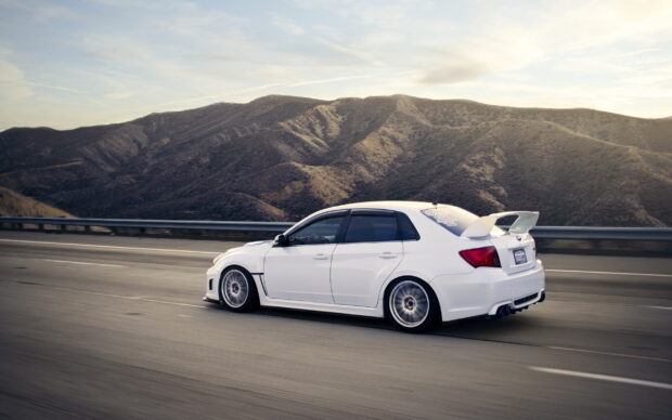A white Subaru WRX is speeding along a highway with mountainous scenery in the background, 2K Desktop Wallpaper