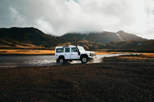 A white off road vehicle is driving through shallow water with mountains in the background, 2K Desktop Wallpaper