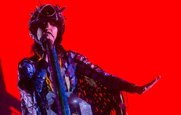 A lead singer from Yeah Yeah Yeahs wearing colorful outfit and helmet sings on stage with red background, HD Desktop Wallpaper
