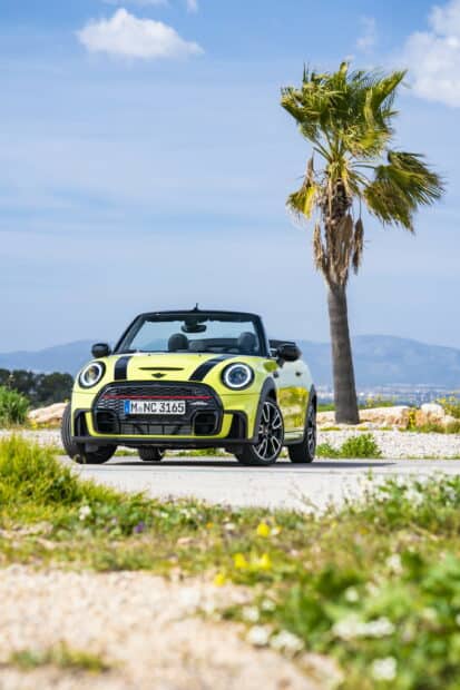 A yellow Mini Cooper with black racing stripes is parked near a palm tree under a blue sky, 2K Phone Wallpaper