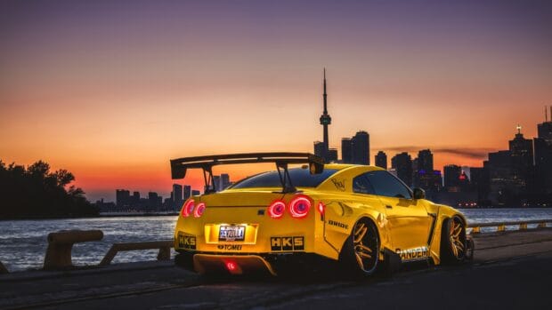 A yellow Nissan GT R is parked beside a river with a city skyline at sunset in the background, 4K Desktop Wallpaper