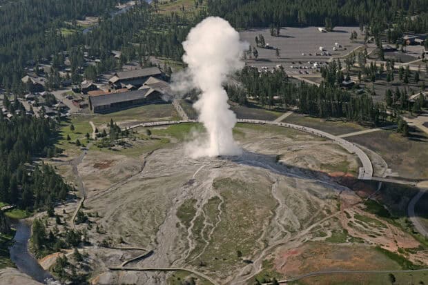 A geyser erupting in Yellowstone National Park with surrounding forest and buildings, HD Desktop Wallpaper