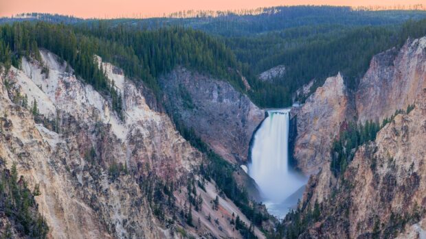 A waterfall flows through rocky cliffs and pine forests in Yellowstone National Park, 4K Desktop Wallpaper