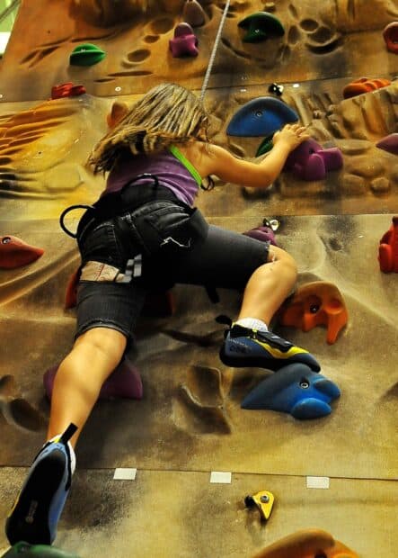 A young girl is climbing an indoor rock wall with colorful holds and safety gear in a gym, HD Mobile Wallpaper