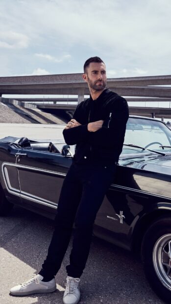 Adam Levine leans on a black Mustang car under overpass wearing a black outfit, 2K Phone Wallpaper