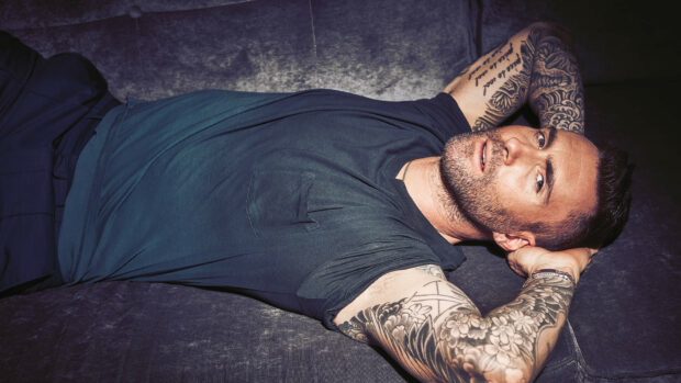 Adam Levine lying on a sofa showing detailed tattoos on his arms, HD Desktop Wallpaper
