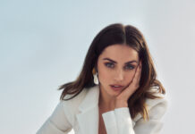 Elegant woman in white blazer and pants, posing outdoors with mountain scenery, showcasing fashion and style.