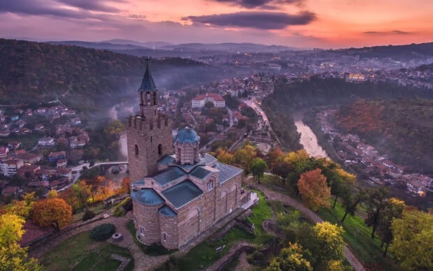 An ancient stone church sits on a hill surrounded by autumn trees and overlooking a Bulgarian town