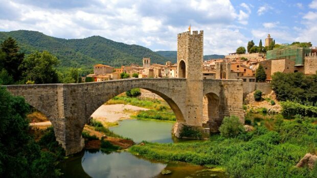 A stone bridge arches over the calm river surrounded by greenery in a historic Spanish town, HD Desktop Wallpaper