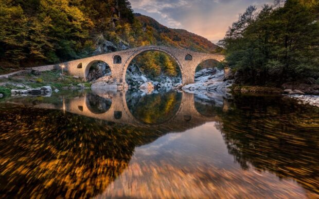 An ancient stone bridge reflects in calm water among autumn trees in Bulgaria, HD Desktop Wallpaper
