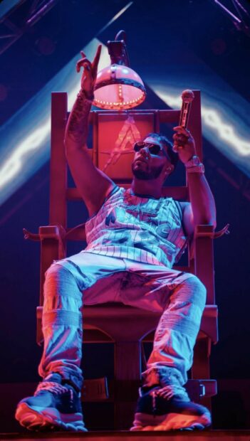 Anuel Aa is sitting in a chair holding a microphone with colorful stage lights, HD Mobile Wallpaper