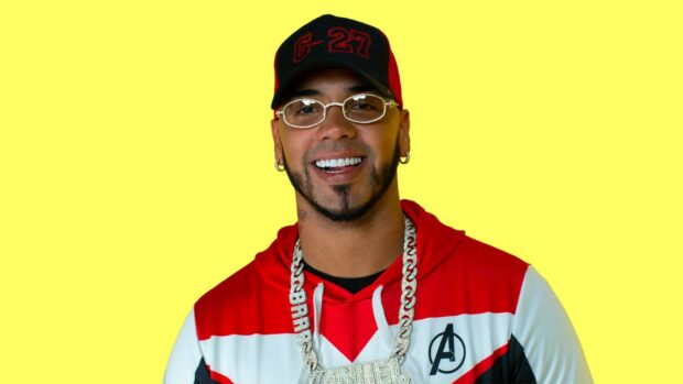 Anuel Aa is wearing a red and white hoodie with a large chain and glasses against a yellow background