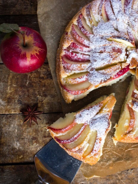 A sliced apple pie dusted with powdered sugar on a wooden surface with a red apple nearby, 2K Desktop Wallpaper