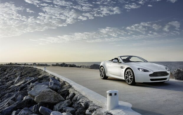 A white Aston Martin convertible car parked on a seaside road with rocky shoreline and cloudy sky