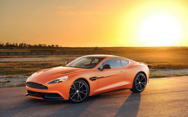 A sleek Aston Martin sports car parked on a road during sunset with an orange sky