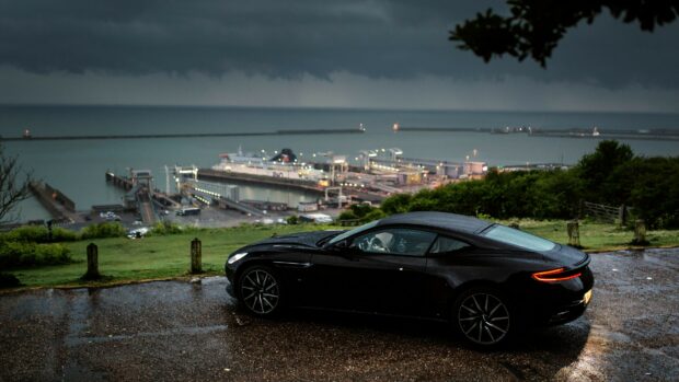 Black Aston Martin parked near a coastal harbor with stormy clouds in the background