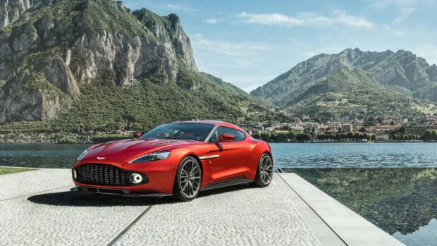 Red Aston Martin sports car parked by lake with mountain landscape in background