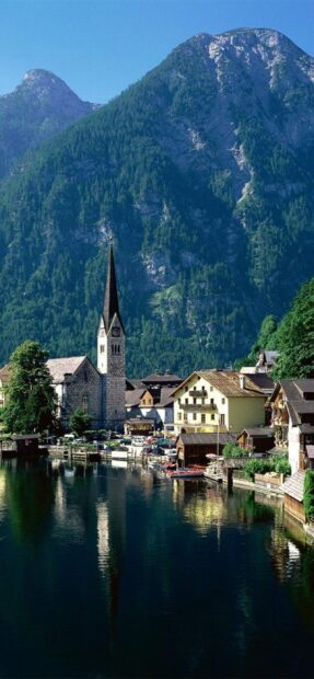 A peaceful Austrian village with a church and houses reflected on a calm lake with mountains behind, 2K Phone Wallpaper