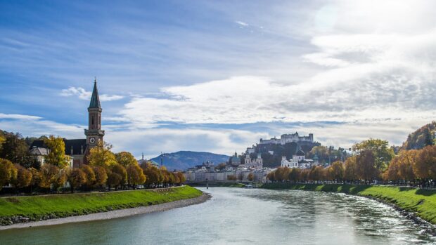 A peaceful river flows through Austria with historic buildings and autumn trees along the banks, 4K Desktop Wallpaper