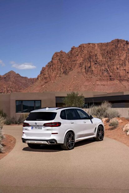 A white BMW X5 driving on a paved road with a mountainous desert landscape and modern house in the background