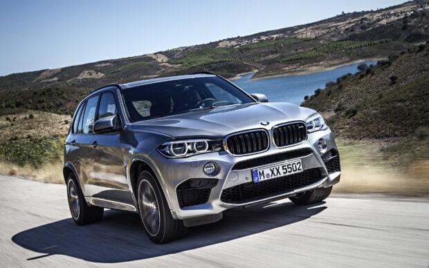 A silver BMW X5 is driving fast on a scenic mountain road near the water, 2K Desktop Wallpaper