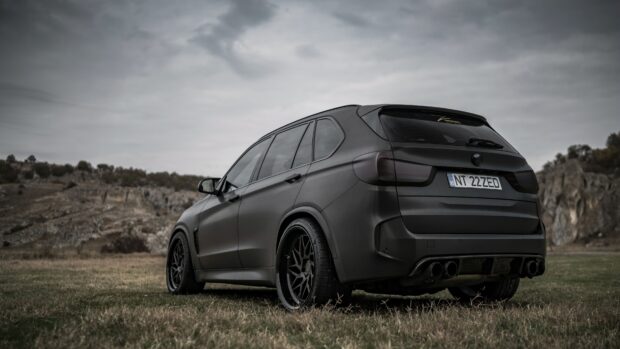 A matte black BMW X5 is parked on grassy terrain with rocky hills under a cloudy sky, 4K Desktop Wallpaper