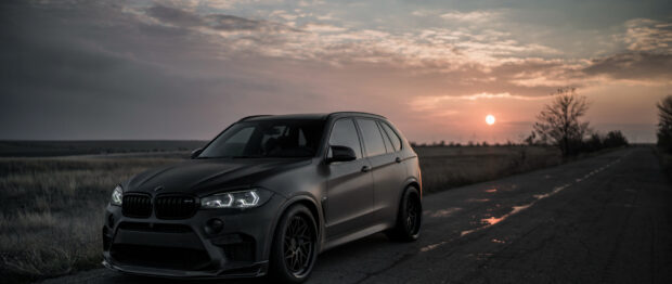 A black BMW X5 is parked on a rural road with a setting sun and cloudy sky in the background, 2K Desktop Wallpaper