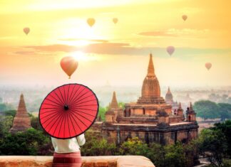 A person holding a red umbrella overlooking Bagan temples and hot air balloons at sunrise