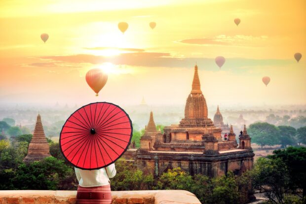 A person holding a red umbrella overlooking Bagan temples and hot air balloons at sunrise