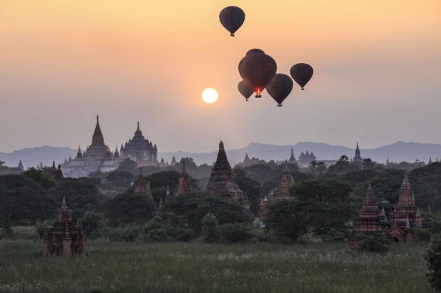 Ancient Bagan temples surrounded by greenery under sunrise with hot air balloons in the sky
