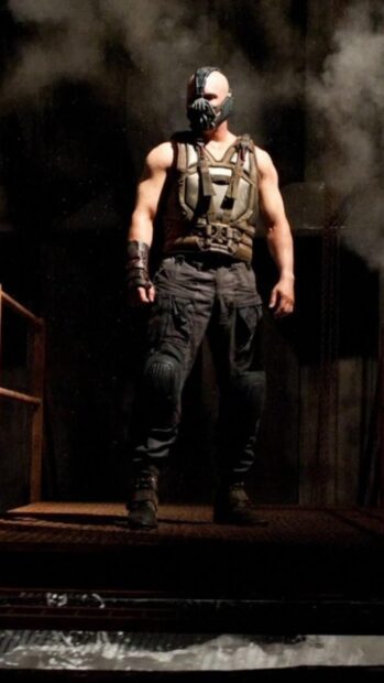 A muscular man wearing a tactical vest and mask representing Bane standing in a dark industrial setting