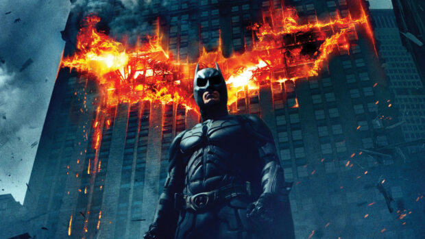 Batman Begins character standing in front of a burning building with a fiery bat symbol in the background