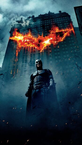 Batman stands in front of a burning building with a fiery bat symbol in the night sky