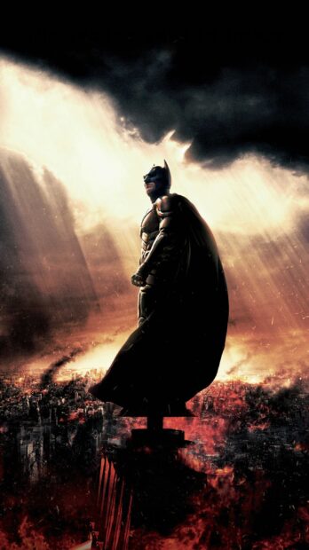 Batman Begins standing on a rooftop overlooking a burning cityscape during a dramatic sunset