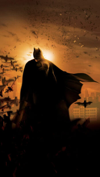 Batman begins character stands with bats flying around at sunset in Gotham city