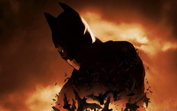 Dark silhouette of Batman with bats flying around against a fiery orange sky in Batman Begins