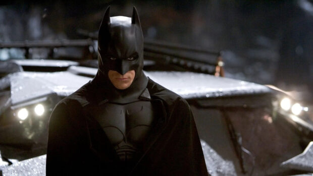 Batman begins character standing seriously in front of the Batmobile in the dark