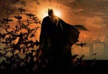 Batman begins character standing with bats flying around in a dark cityscape at sunset